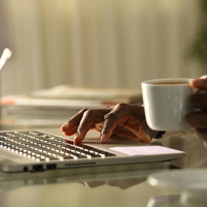 Foto einer Person, die mit einer Kaffetasse in der Hand an einem Notebook arbeitet.